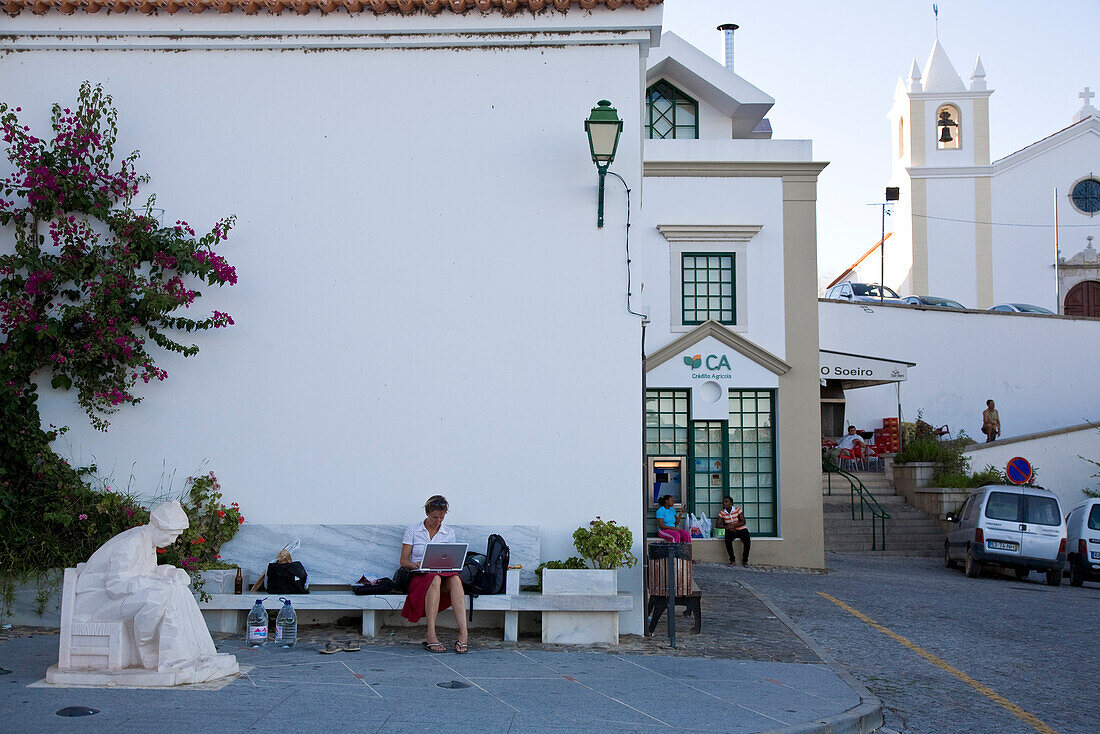 Yong women using her laptop, notebook, using free internet access, W-LAN, white church and village square, Alcoutim, Algarve, Portugal