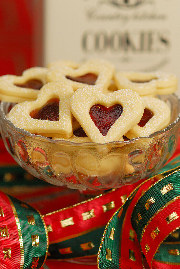 Weihnachtsgebäck Himbeerherzen mit Puderzucker bestäubt türmen sich in Glasschale, Weihnachtsdekoration im Hintergrund