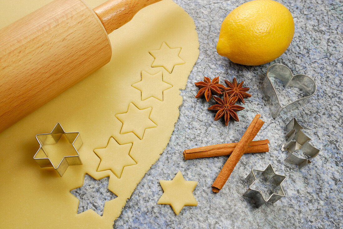 Stars cutted out of cookies dough, cookie cutters for Christmas cookies, rolling pin, lemon, cinnamon and star anise laying at worktop
