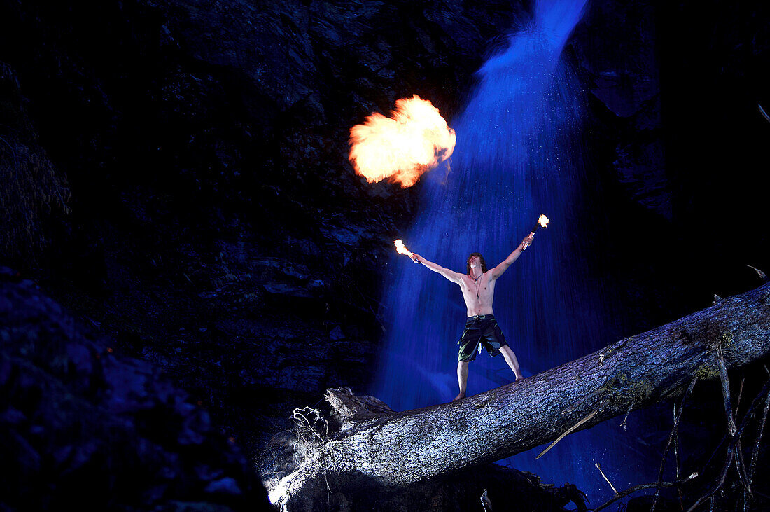 Junger Mann steht auf einem Baumstamm vor einem Wasserfall, spuckt Feuer, See, Tirol, Österreich