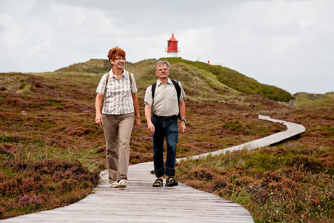 Couple walking along wooden path through dunes, lighthouse in background, Amrum island, Schleswig-Holstein, Germany