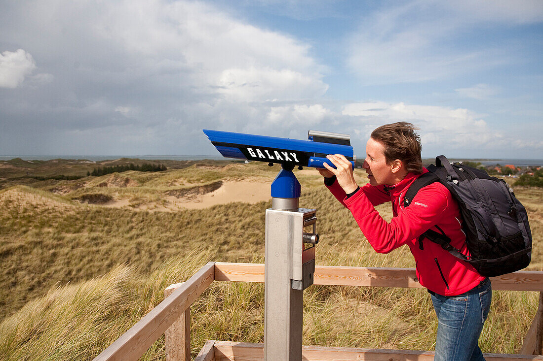 Woman looking through coin-operated binoculars over dunes, Norddorf, Amrum island, Schleswig-Holstein, Germany