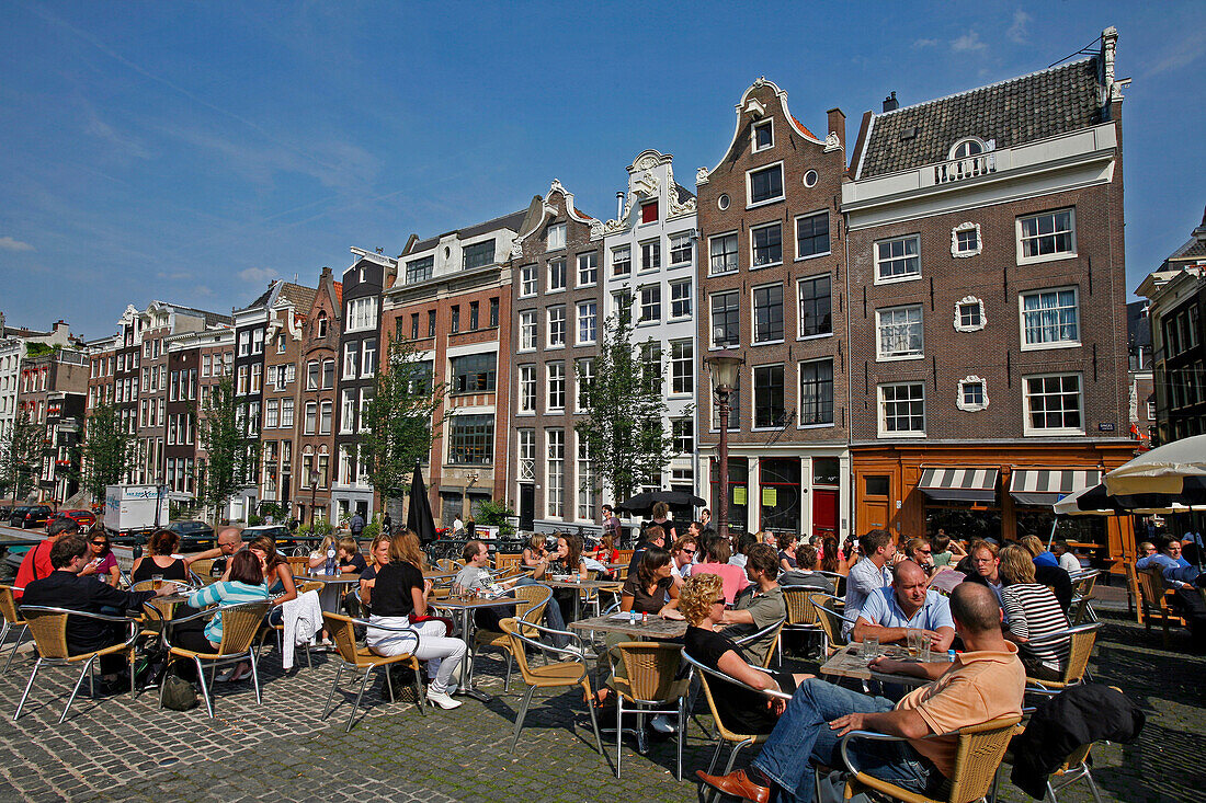 Group Of People Sitting At A Sidewalk Cafe By A Canal And The Facade Of A Traditional House In The Background