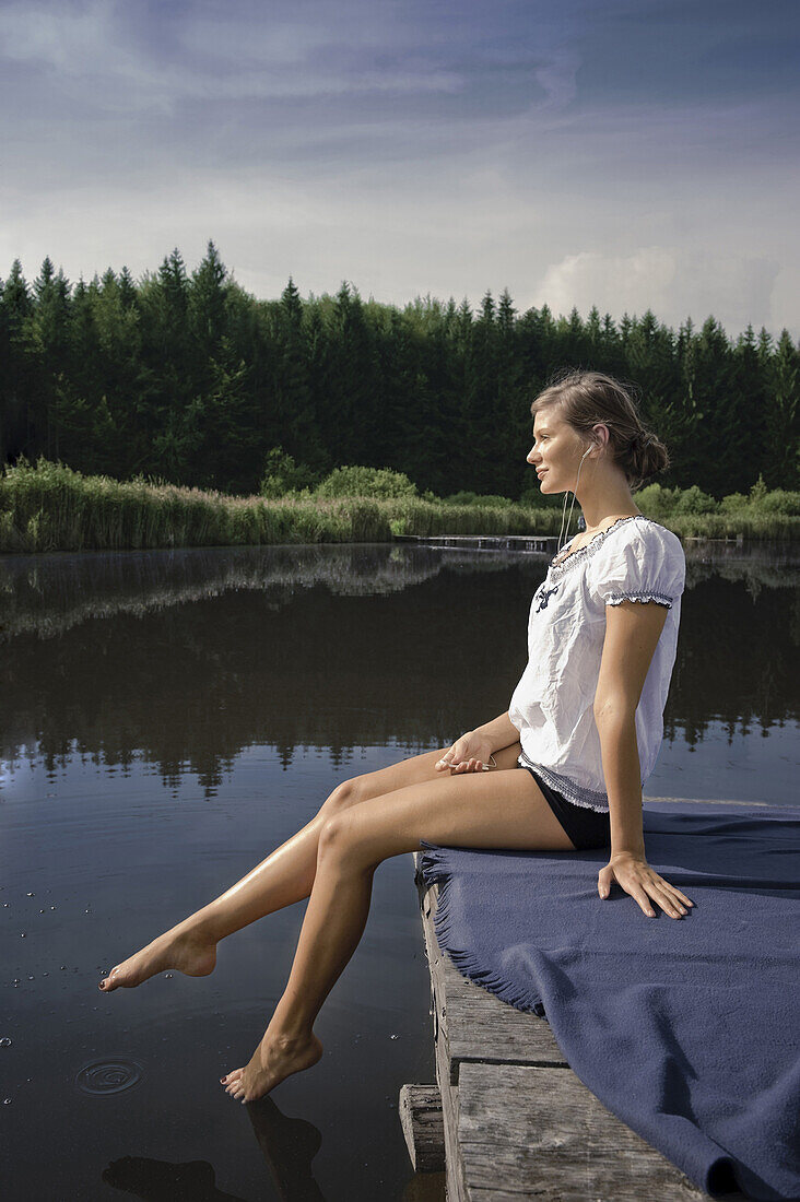 Junge Frau sitzt auf einem Steg am Starnberger See, hört Musik, Bayern, Deutschland