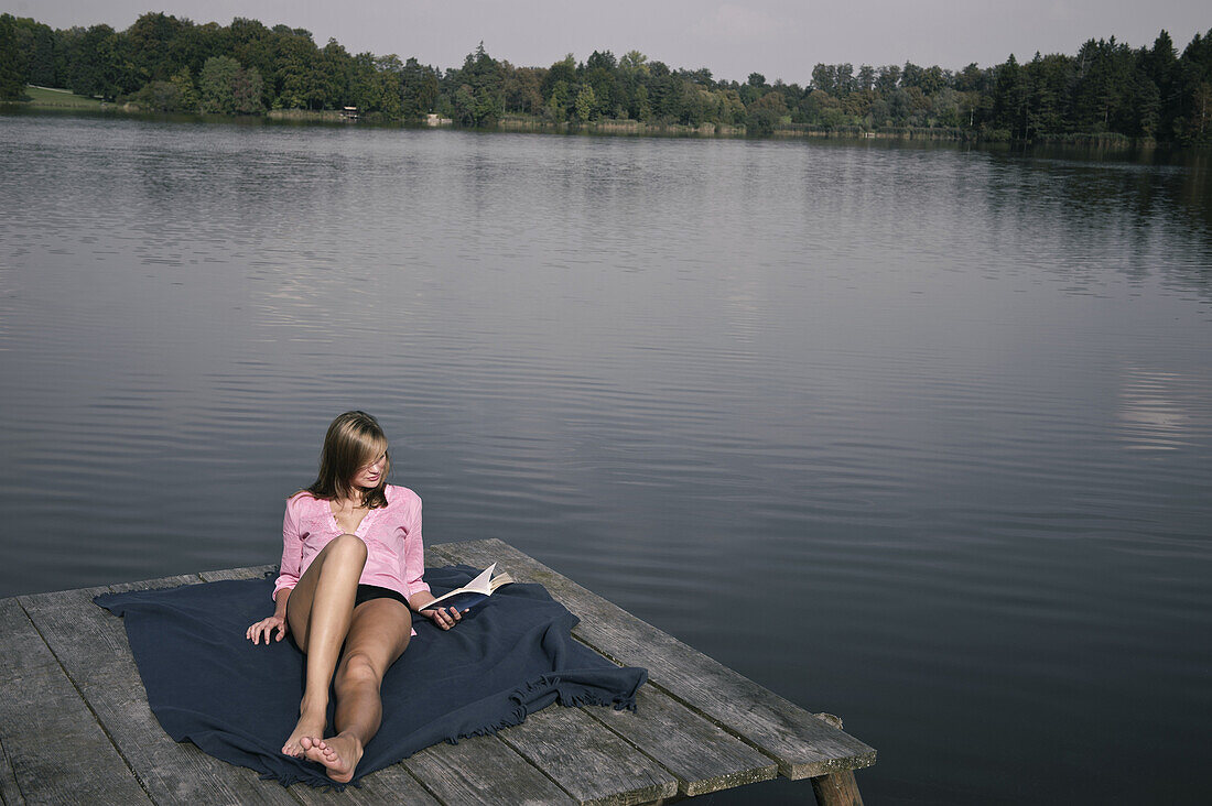 Junge Frau liegt auf einem Steg und liest ein Buch, Starnberger See, Bayern, Deutschland