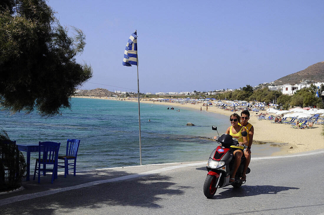 Junges Paar auf Motorroller vor Strand im Sonnenlicht, Insel Naxos, Kykladen, Griechenland, Europa