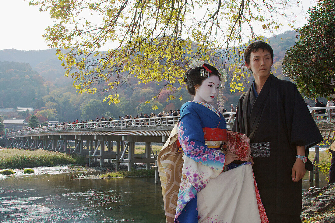 A Japanese man wearing a traditional kimono and a woman dressed up as a geisha are standing infront of the famous bridge Togetsu-kyo,  the dominant landmark in Arashiyama with autumn leaf colors in the back ground