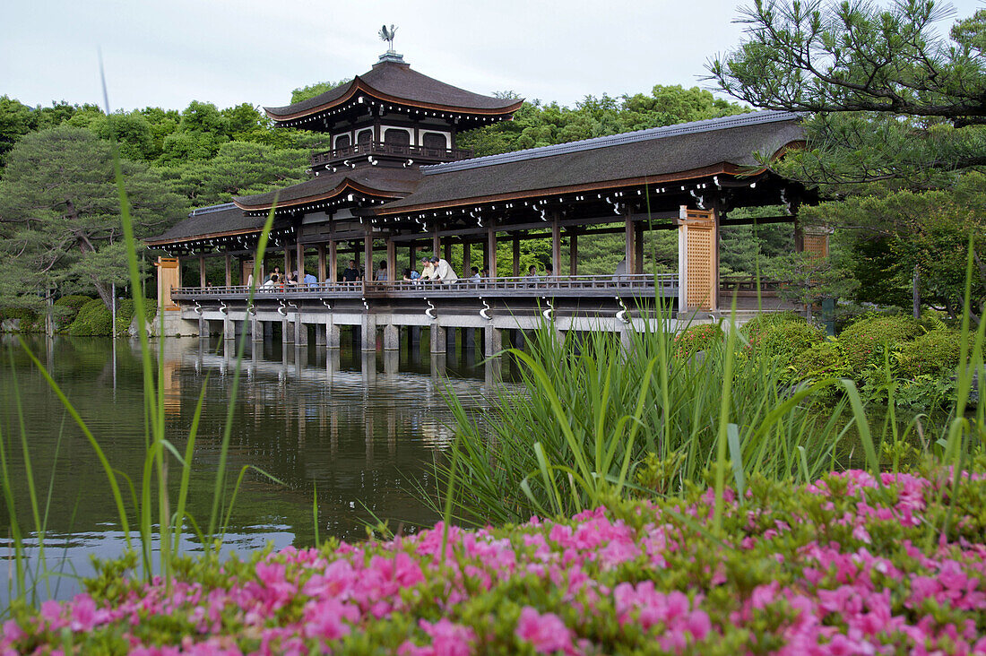 A Chinese inspired covered bridge over a pond at Heian shrine,  with pink flowers and green grasses in the foreground in Kyoto city.