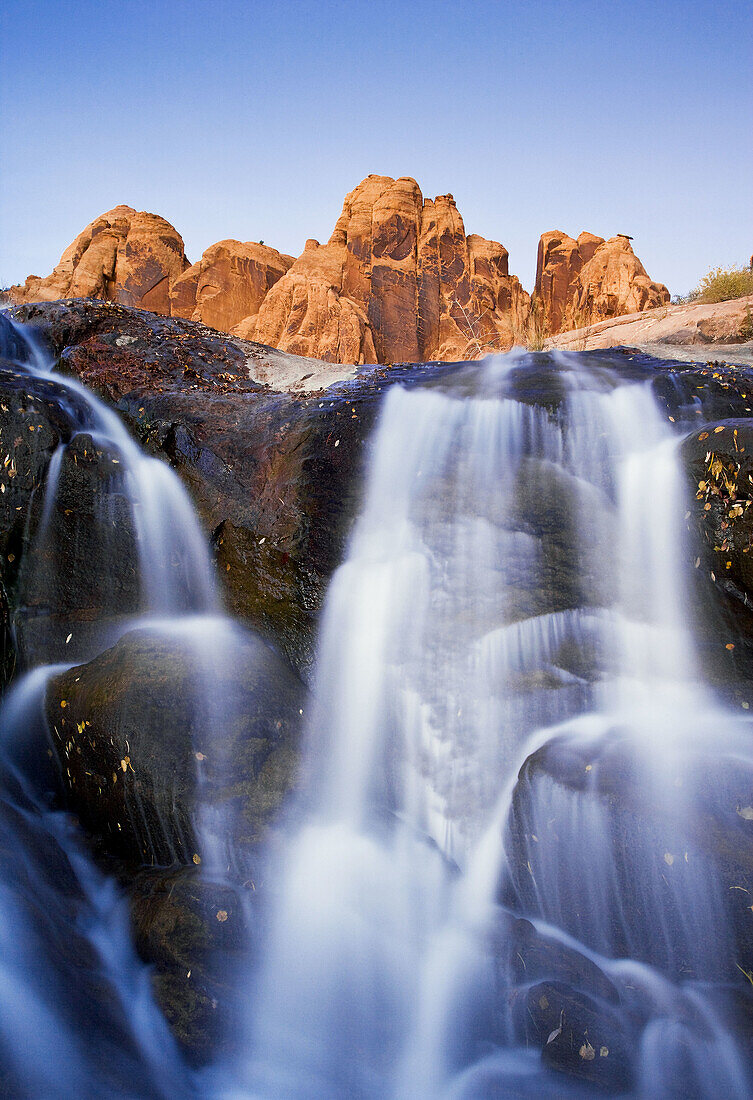 A waterfall tumbles over rocks below a jagged sandstone ridge illuminated in the last light of the day near Moab,  Utah
