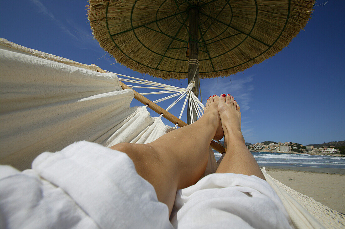Womans' legs ona  hammock under thatched a sun shade, Mallorca, Balearic Islands, Spain