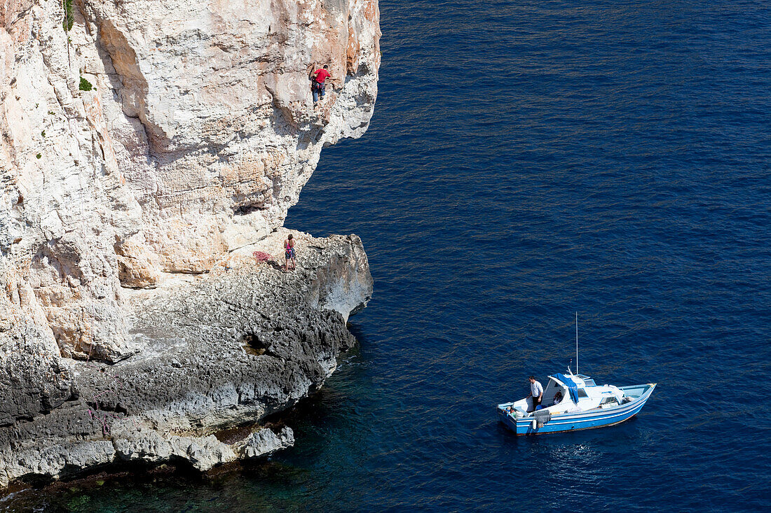 A young woman and a young man climbing on the cliffs at the bay of Zurrieq, a little boat closeby, Malta, Europe