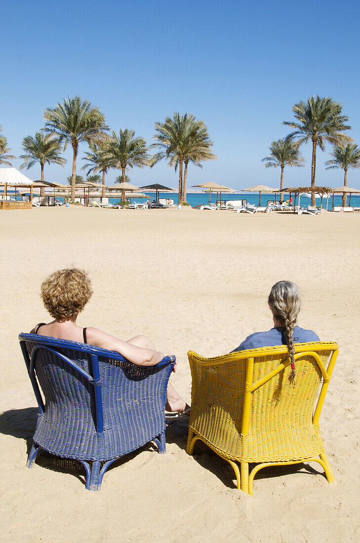 Two women sitting on wicker armchairs on beach and palm trees and Red sea at Hurgadah, Egypt