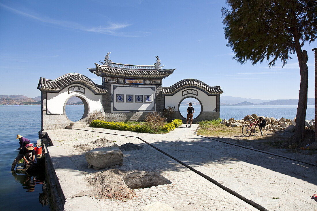 Mondtor am Erhai See, traditionelle chinesische Architektur, Touristin mit Fahrrad, Frauen waschen die Wäsche im See, Yunnan, Volksrepublik China, Asien