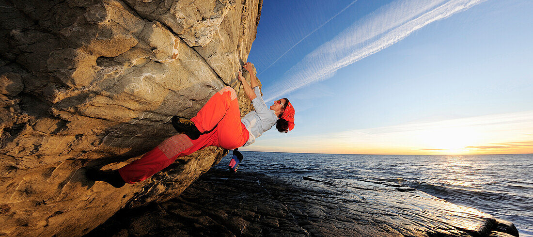Panorama with climber, woman bouldering at an overhanging rock along the Mediterranean coast at sunset, Mediterranean in the background, Liguria, Italy