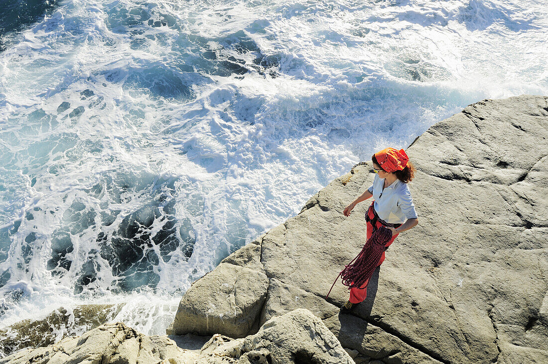 Frau hält Kletterseil in der Hand und blickt auf gischtendes Meer, Steilküste am Mittelmeer, Ligurien, Italien