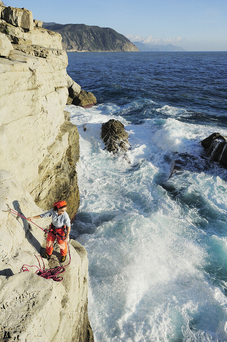 Woman standing on a cliff and rappelling, abseiling towards the spraying sea, cliff at the Mediterranean coast, Liguria, Italy