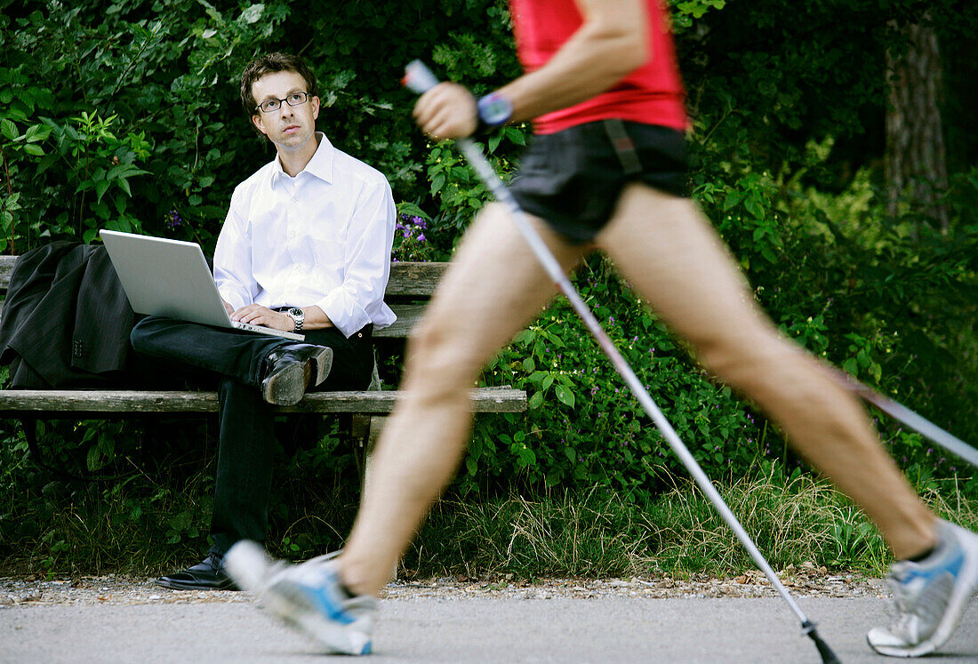 Geschäftsmann mit Laptop auf einer Parkbank, Nordic Walker läuft vorbei