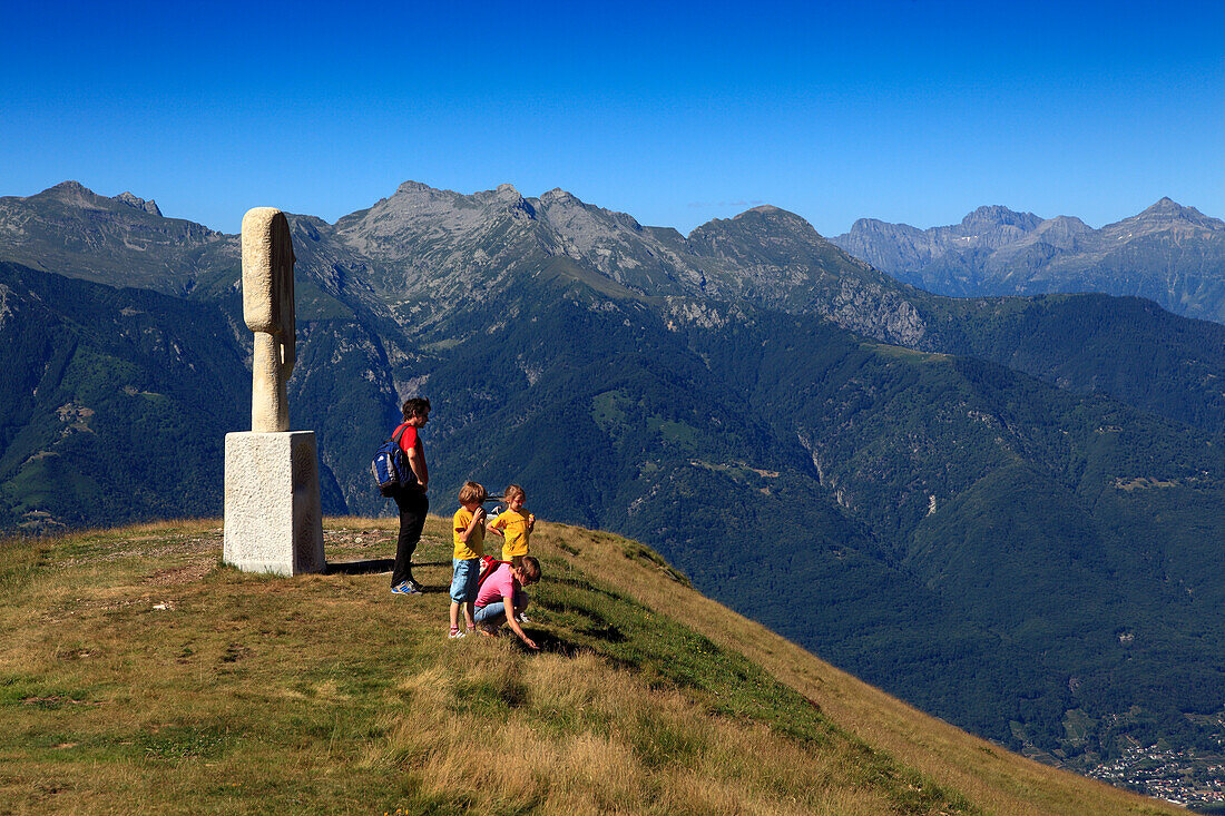 Couple with kids in front of a sculpture on Alpe Foppa, hike in the mountains to Monte Tamaro, Ticino, Switzerland