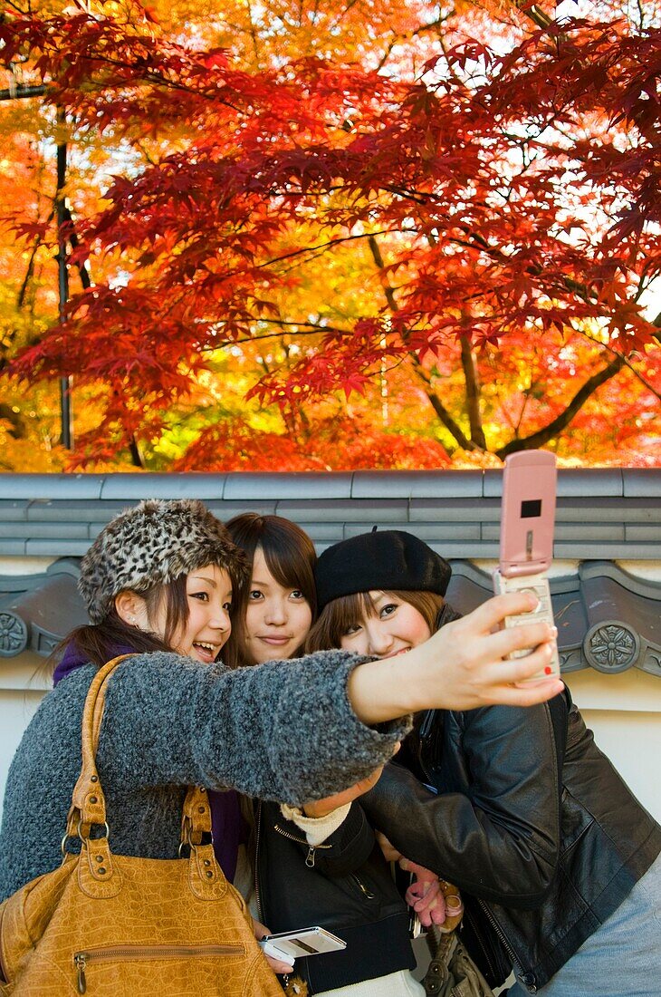 Three women take a souvenir photograph of themselves in front of colourful autumn leaves in Kyoto Japan