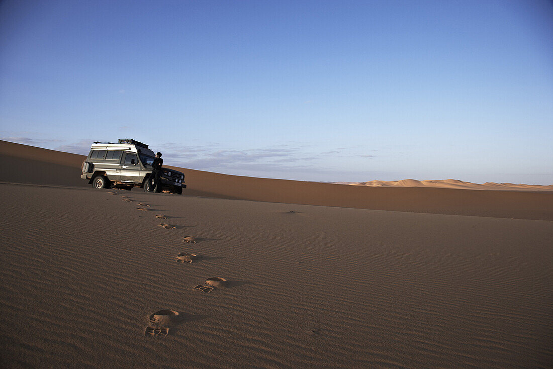 Foot prints in the sand, woman standing in front of a Toyota Landcruiser, Murzuk sand sea, Lybia, Africa