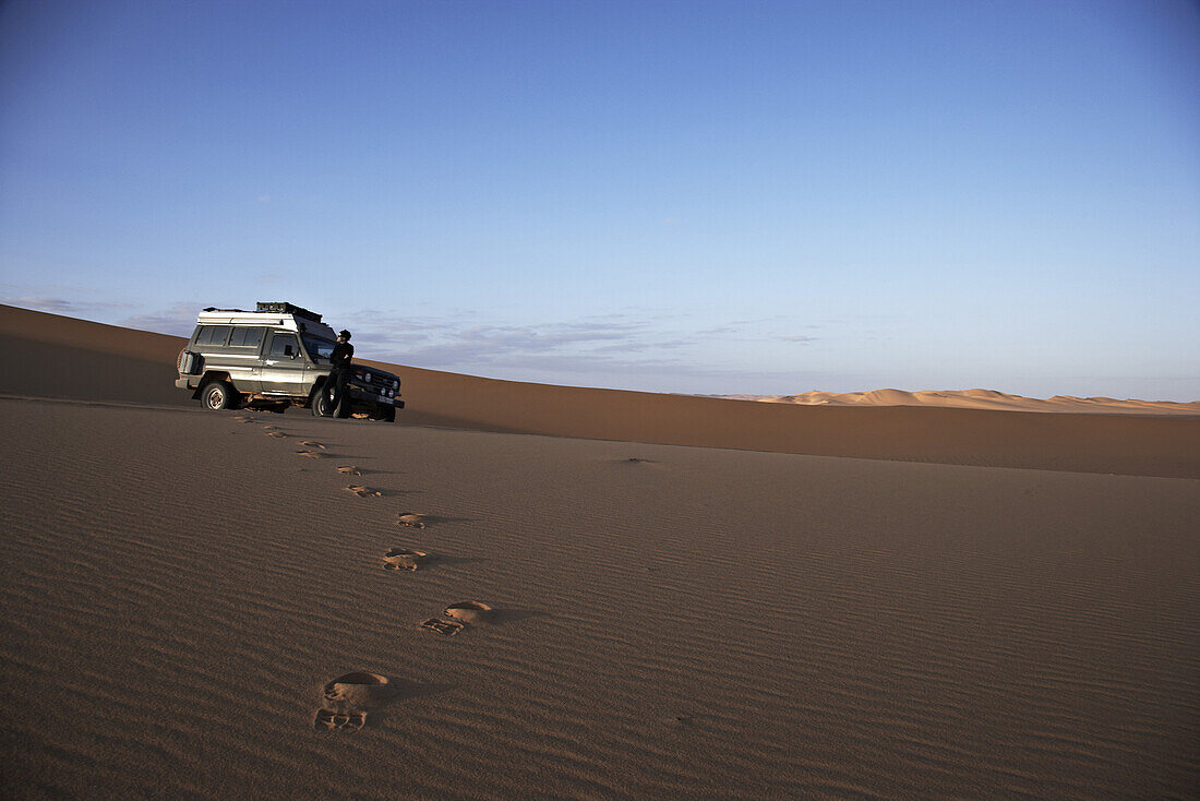Fußspuren im Sand, Frau steht vor Toyota Landcruiser, Murzuk Sandmeer, Libyen, Afrika