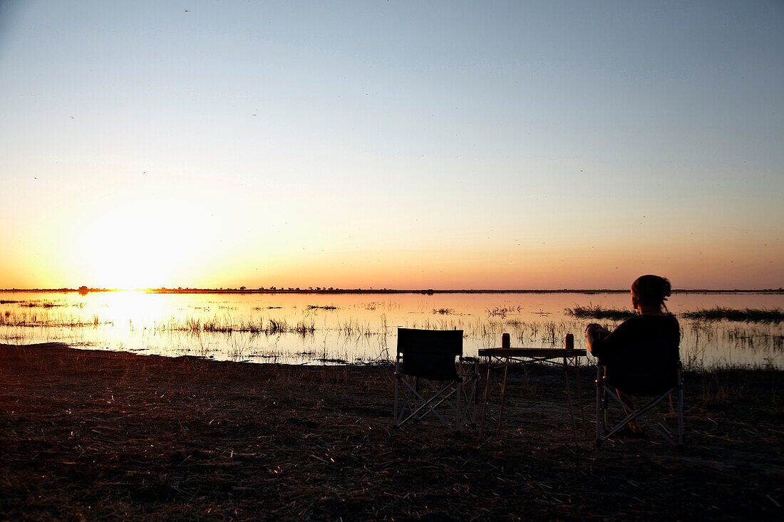 Frau sitzt auf Campingstuhl und blickt in Sonnenuntergang, Chobe Nationalpark, Botswana, Afrika