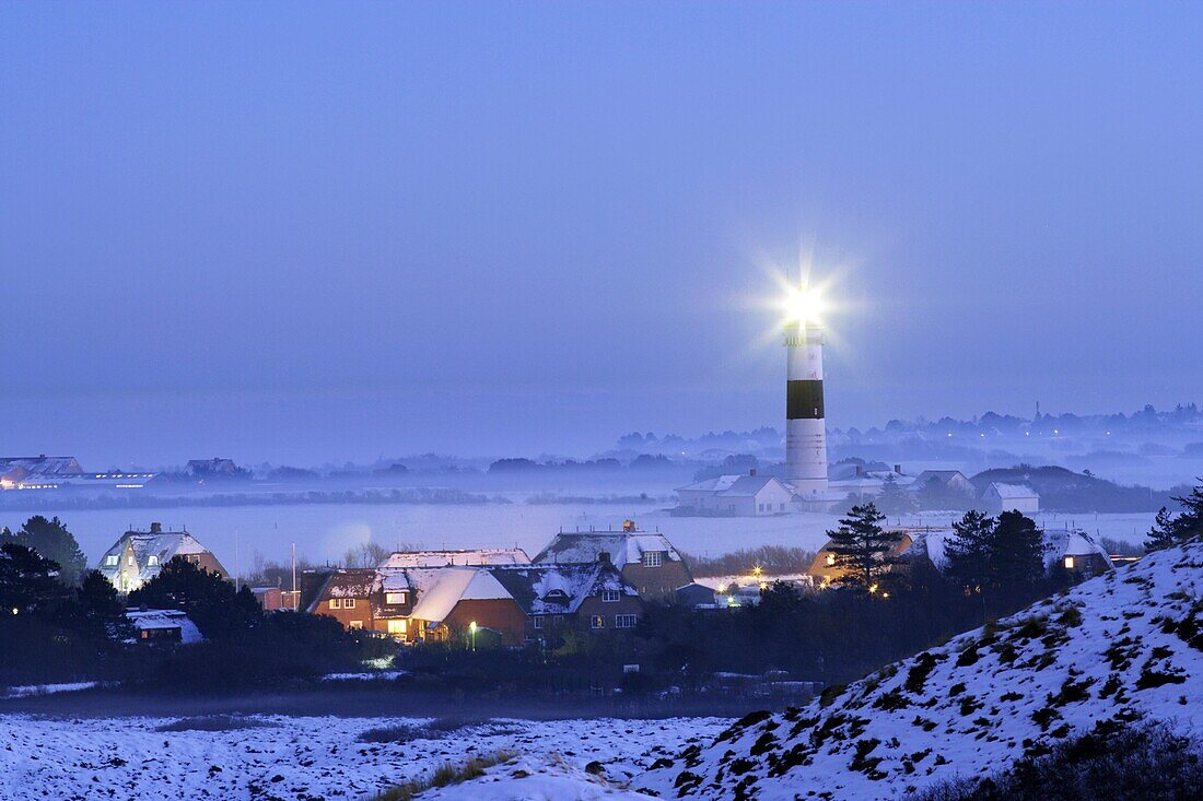 Lighthouse Kampen on snow covered fields in operation with the light beam visible, seen from abvoe with the traditional houses of the village of Kampen and a bright blue sky of the early evening, Sylt, Northfrisian Islands, Schleswig-Holstein, Northern Ge