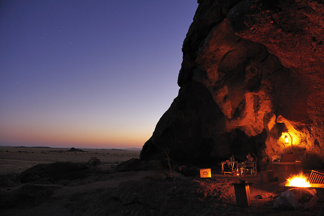 Personen sitzen bei Grillfeuer unter Felsüberhang, Namib Naukluft National Park, Namibwüste, Namib, Namibia