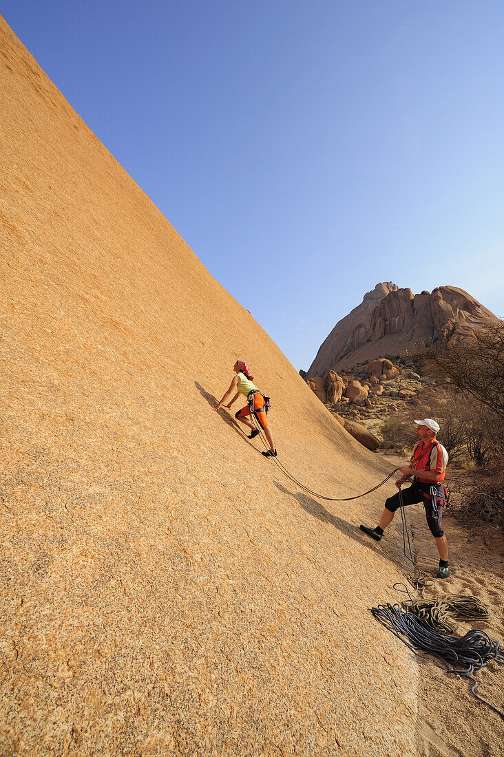 Frau klettert an roter Felswand und Mann sichert, Große Spitzkoppe, Namibia