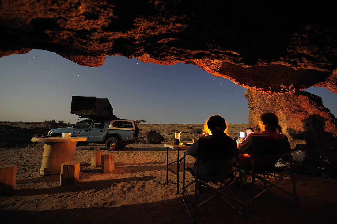 Gruppe Personen sitzt unter Felsüberhang, Auto mit Dachzelt im Hintergrund, Felswüste, Namib Naukluft National Park, Namibwüste, Namib, Namibia