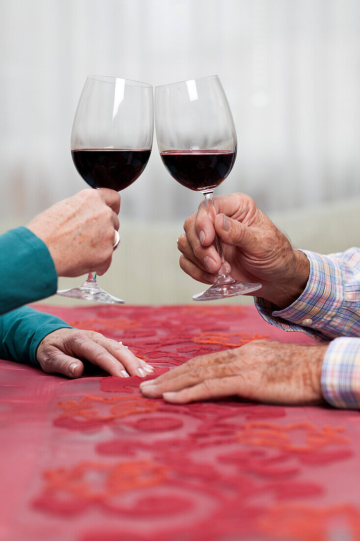 Hände von zwei alten Menschen, sie prosten sich mit einem Glas Rotwein zu