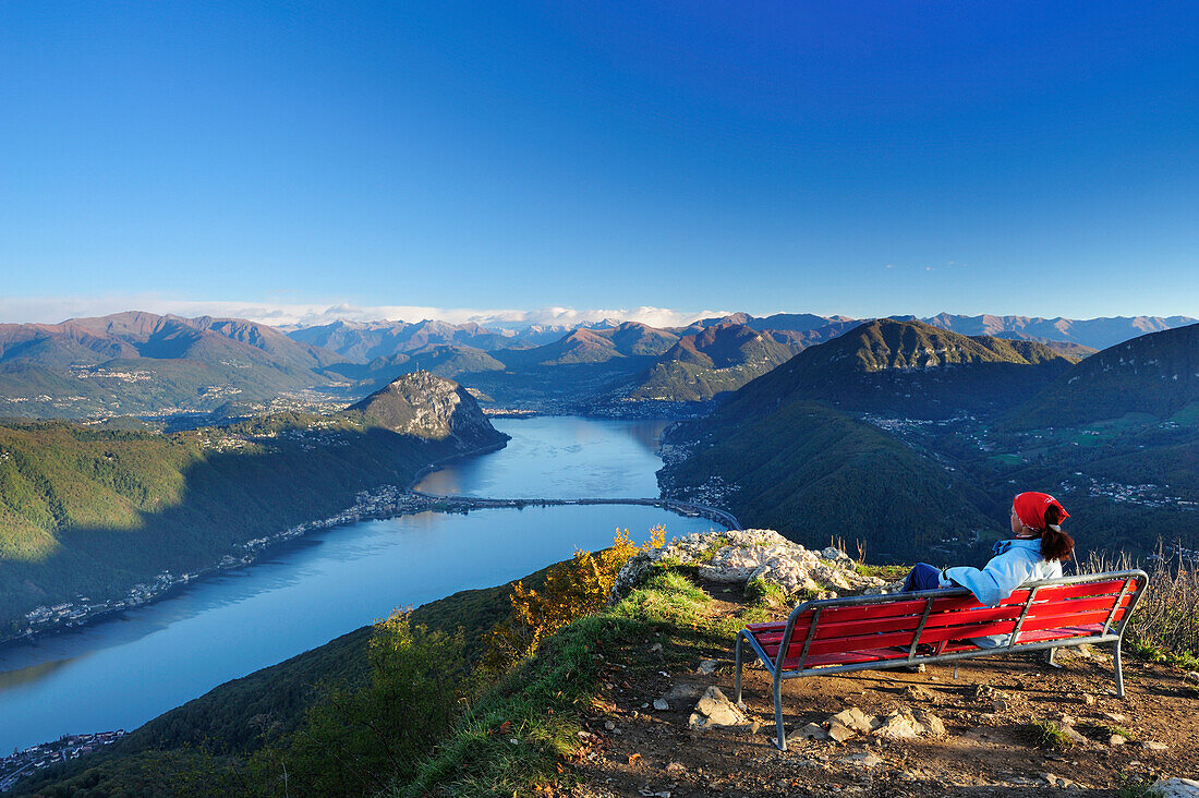 Woman sitting on red bench and looking towards lake Lugano, Ticino range in background, View from the  Monte San Giorgio, UNESCO World Heritage Site Monte San Giorgio, lake Lugano, Ticino range, Ticino, Switzerland, Europe
