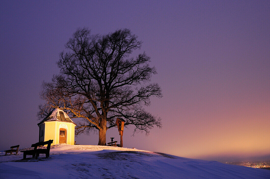 Illuminated chapel in front of cross and oak tree in winter, Chiemgau, Upper Bavaria, Bavaria, Germany, Europe