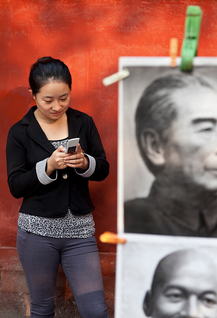 Young women with mobile, cellphone, in front of s red wall, picture of Zhou Enlai, former prime minister of China, book market in Ditan Park in Beijing, Beijing, People's Republic of China
