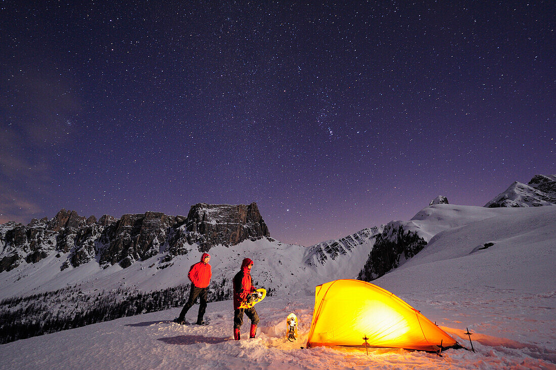 Two persons with snow shoes in front of illuminated tent on snow face in front of Croda da Lago and Monte Formin, Passo Giau, Cortina d' Ampezzo, UNESCO World Heritage Site Dolomites, Dolomites, Venetia, Italy, Europe