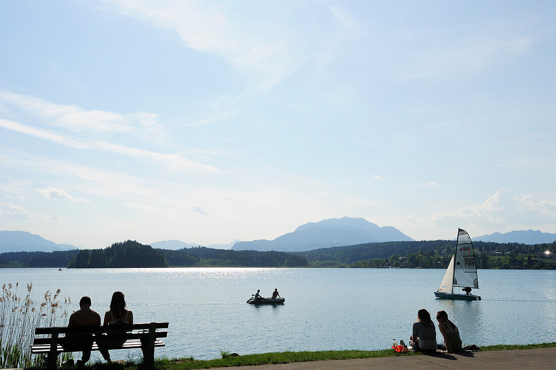 Bathers sitting at lakeside of lake Faaker See with rowing boat and sailing boat, lake Faaker See, Carinthia, Austria, Europe