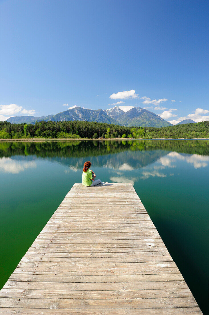 Frau sitzt auf Holzsteg am Turnersee und blickt auf Karawanken, Turnersee, Kärnten, Österreich, Europa
