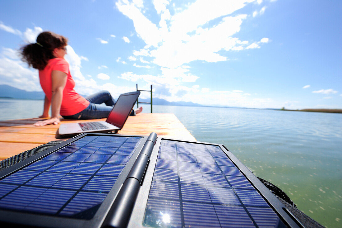 Woman sitting on a jetty at lake Chiemsee, laptop and solar panel in foreground, Chiemgau, Upper Bavaria, Bavaria, Germany