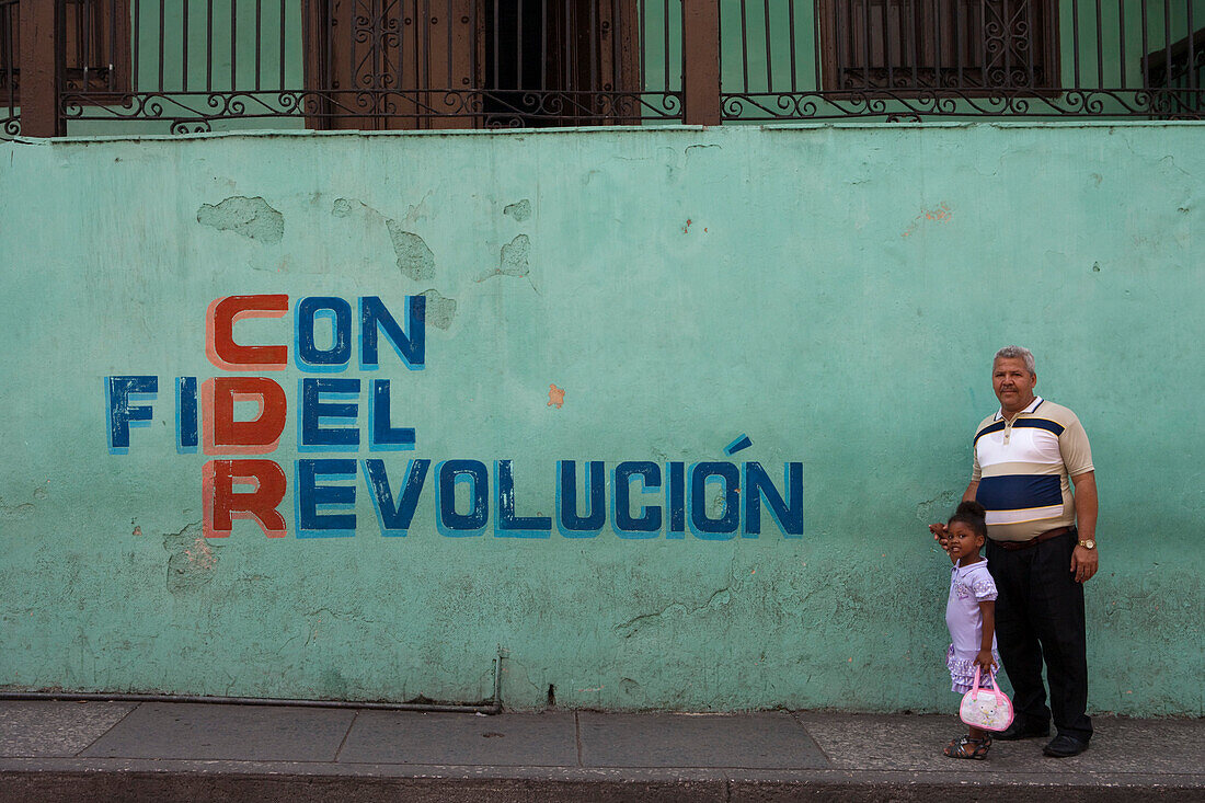 Vater und Sohn vor Wandgemälde mit Propaganda Schriftzug, Santiago de Cuba, Kuba, Karibik