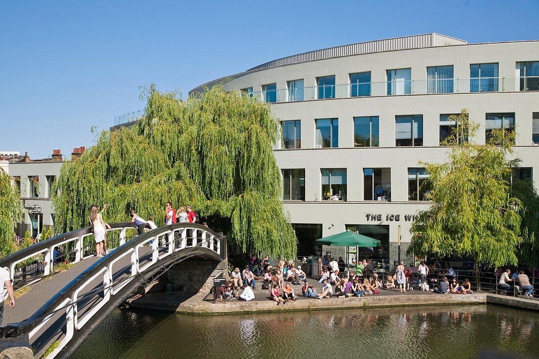 People sitting in sun by canal outside The Ice Wharf, Camden Lock, London, England, UK