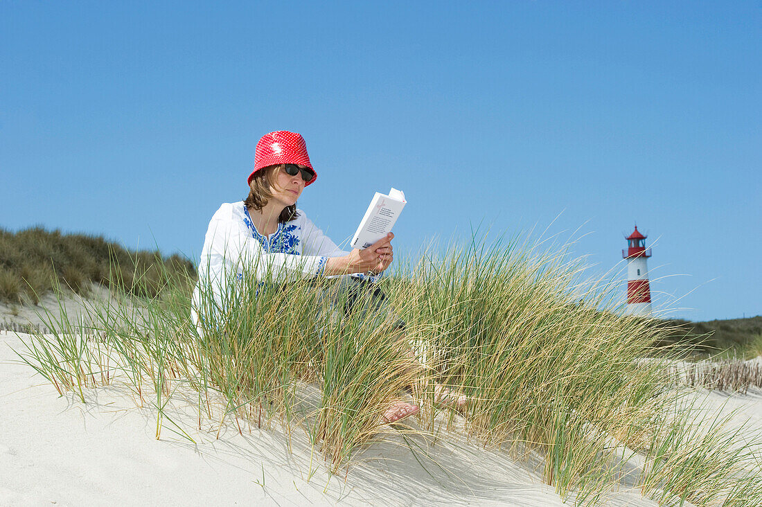 Frau mit Hut sitzt am Strand und liest, Leuchtturm List-Ost im Hintergrund, Ellenbogen, List, Sylt, Schleswig-Holstein, Deutschland
