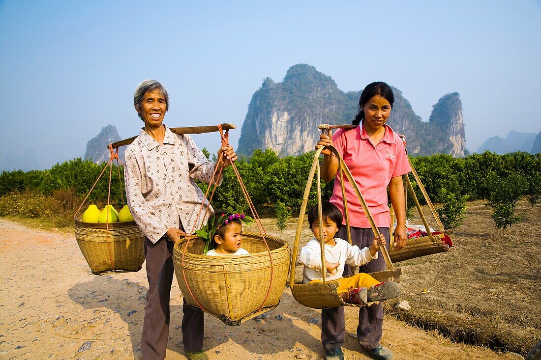 China, Guangxi Zhuang Autonomous Region, Yangshuo County Two rural farm workers carry fruit and their children in hand woven baskets along a rural road running through an agriculture landscape dominated by the Karst peaks of Yangshuo County