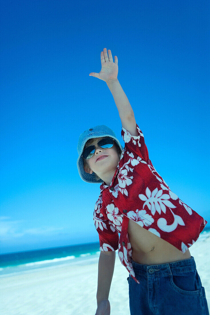 Boy standing on beach with hand raised, wearing beach clothes