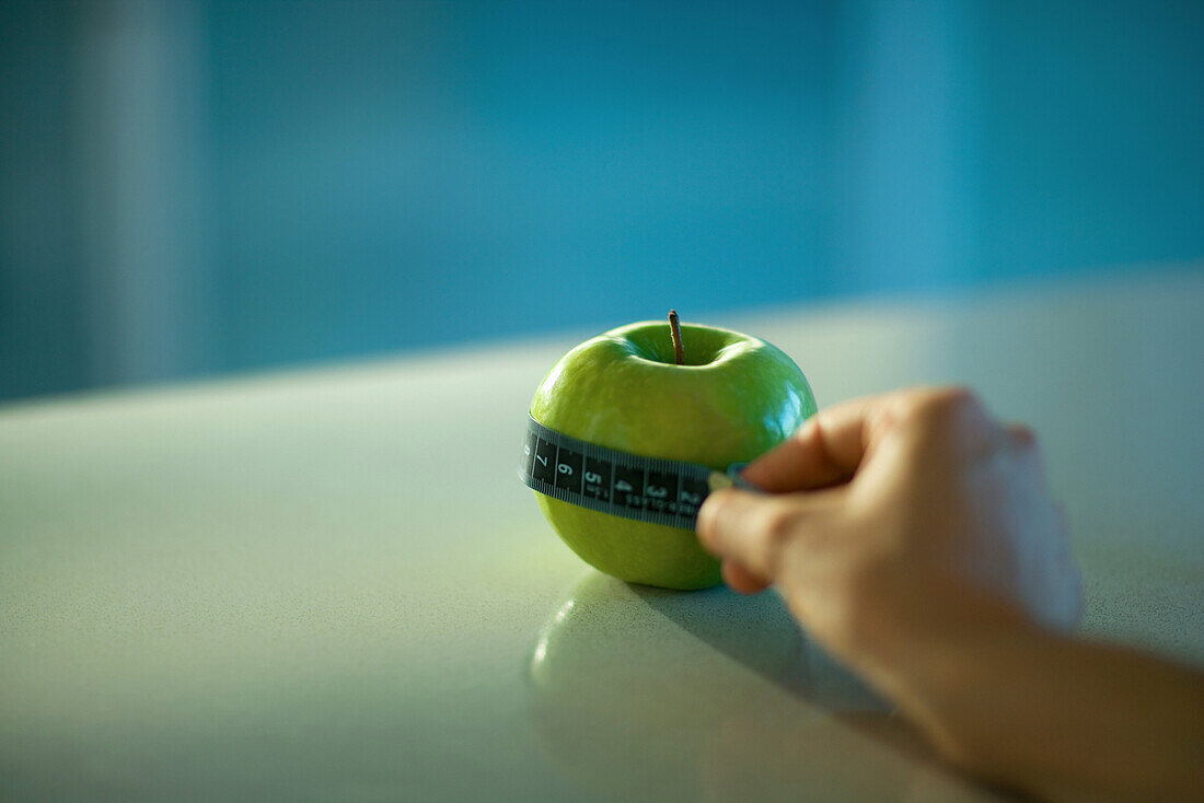 Woman holding measuring tape around apple, cropped view of hand