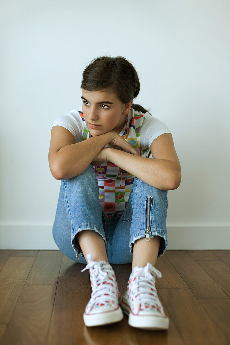 Teenage girl sitting on hardwood floor, hugging knees, looking away from camera