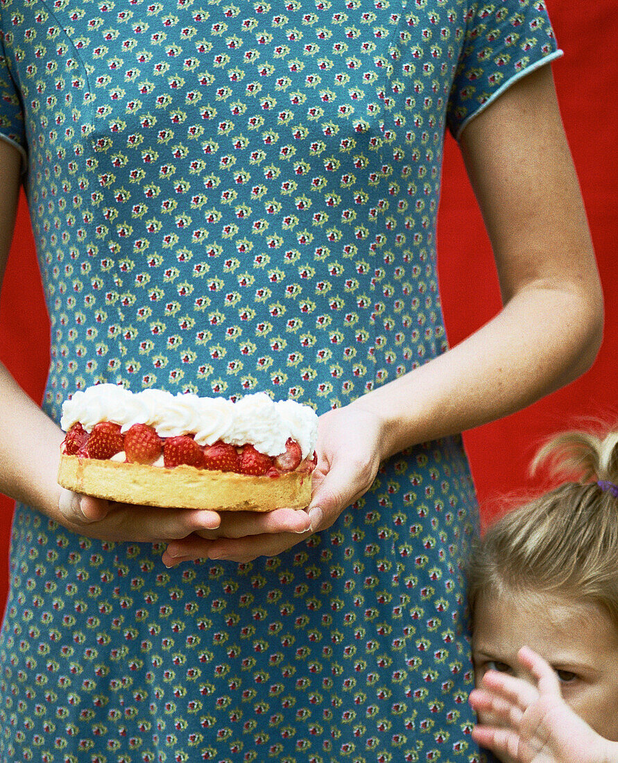 Woman holding cake, mid section, and child covering face with hand