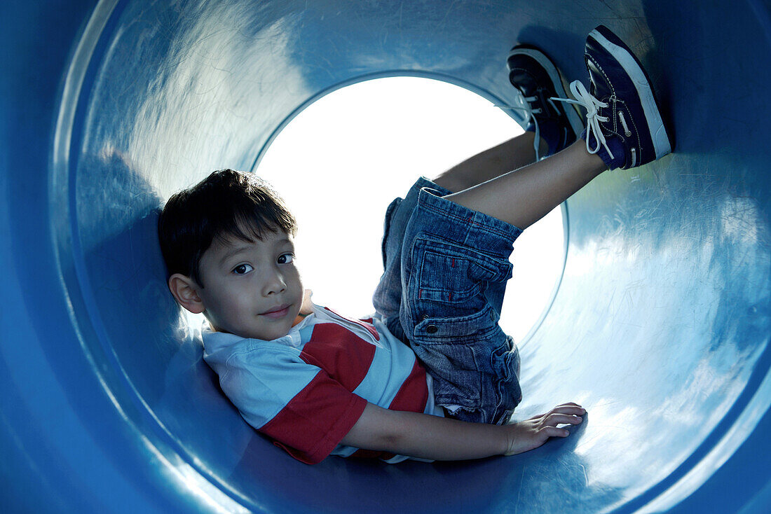 Boy sitting in playground tunnel with legs up, looking at camera, full length