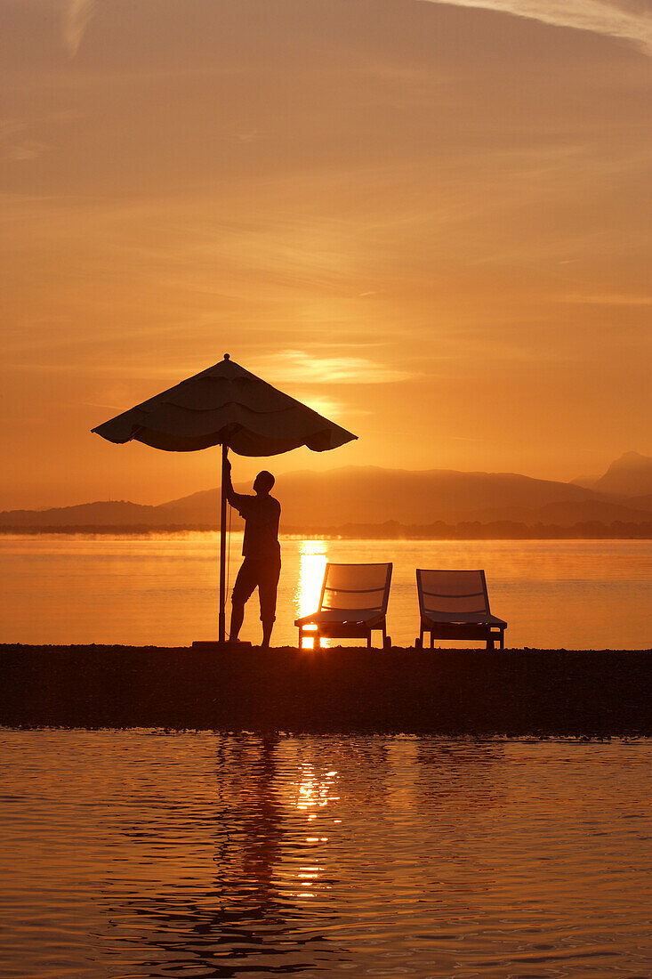 Sonnenaufgang Krautinsel, Mann öffnet Sonnenschirm, Chiemsee, Bayern, Deutschland