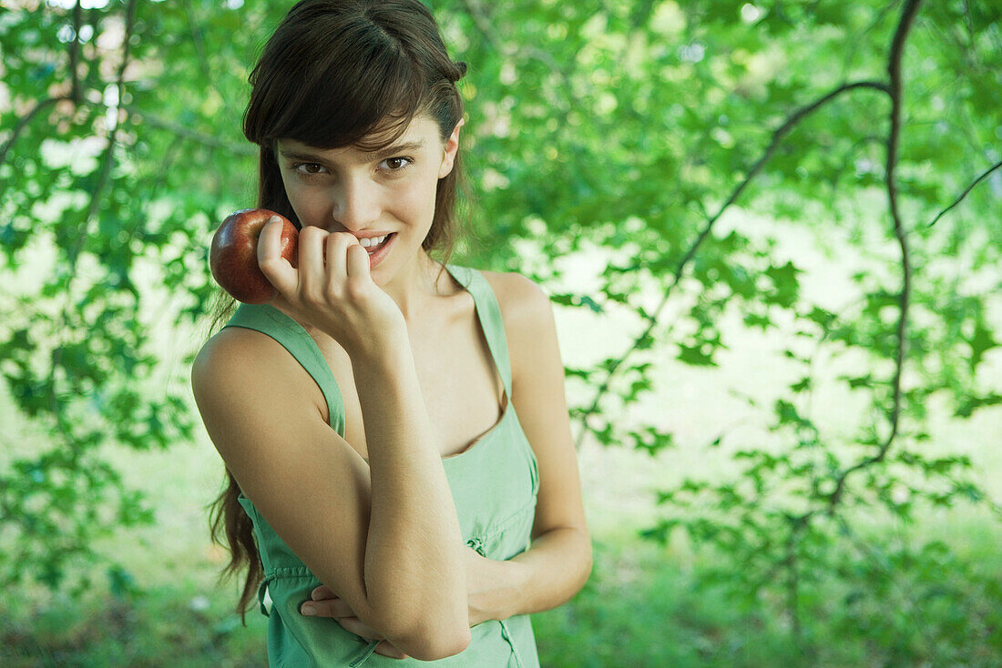 Junge Frau hält im Freien einen Apfel hoch und lächelt in die Kamera
