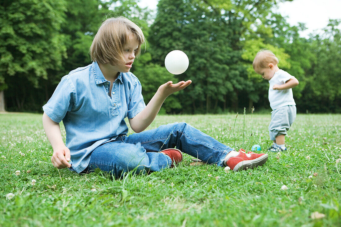 Girl with Down's Syndrome sitting on the ground, throwing ball in the air, baby boy standing in background